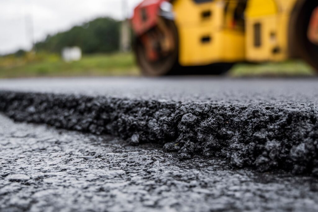 A close up view of asphalt that was used to pave a road with a roller being used in the background